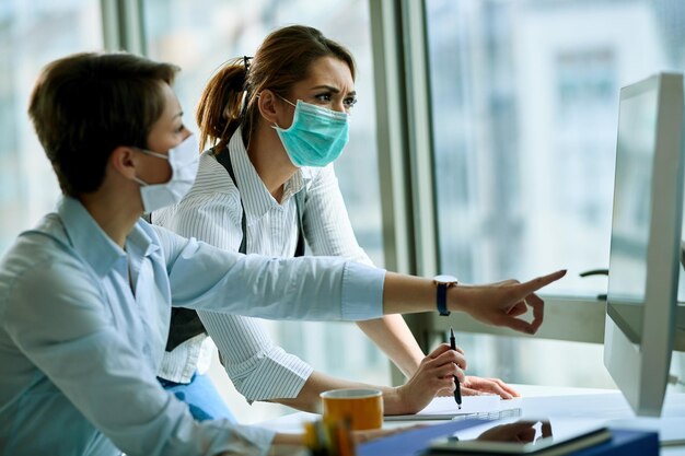 Young businesswoman and her colleague wearing face masks while working on a computer in the office during virus epidemic