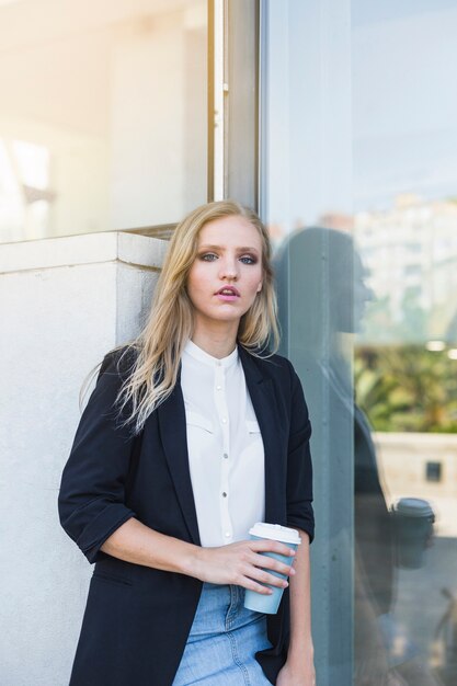 Young businesswoman leaning on wall holding take away coffee cup