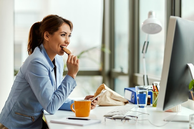 Young businesswoman using cell phone while eating a cookie at her office desk