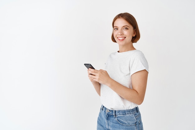 Free Photo young candid girl using smartphone and smiling . woman chatting on mobile phone app, standing against white wall