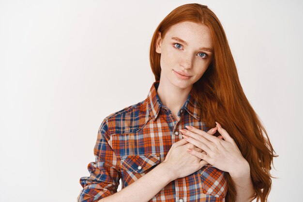 Young caring and heartfelt woman holding hands on heart, thanking for something, standing over white wall