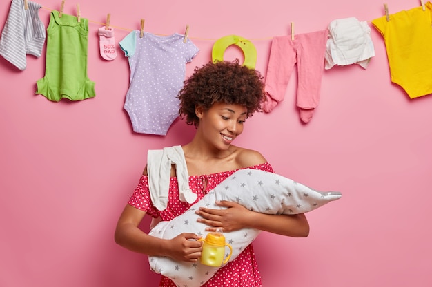 Young caring mother cares about newborn child, feeds with milk, enjoys happy moments of motherhood, poses at home. Little baby on artificial feeding. Babysitting, parenting concept. Child birth