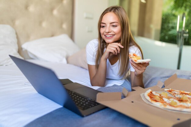 Young casual woman lying on a bed in bed at home while eating pizza and using laptop