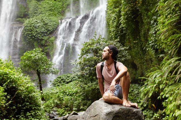 Young Caucasian barefooted male tourist with backpack sitting on rock surrounded by rainforest and admiring gorgeous view with waterfall