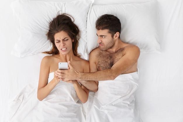 Young Caucasian couple fighting in bed: unshaven man trying to snatch mobile phone from his wife's hands