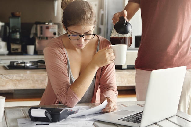 Free photo young caucasian couple having financial problems. stressed female in glasses drinking coffee while managing family budget, sitting at kitchen table with documents, notebook computer and calculator