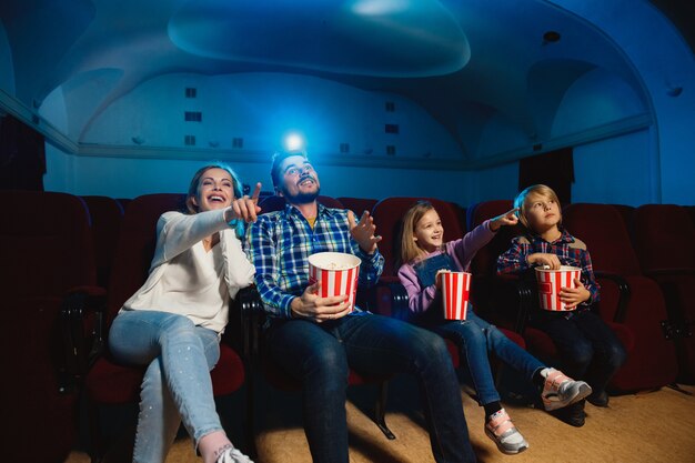 Young caucasian family watching a film at a movie theater, house or cinema. Look expressive, astonished and emotional. Sitting alone and having fun. Relation, love, family, childhood, weekend time.