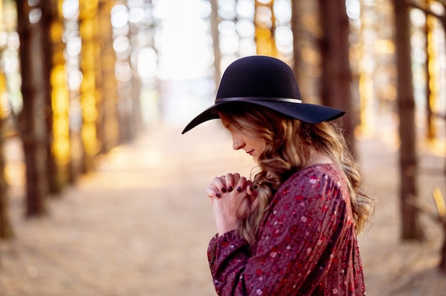 Young Caucasian female in a stylish black hat praying in the scenic forest, autumn mood