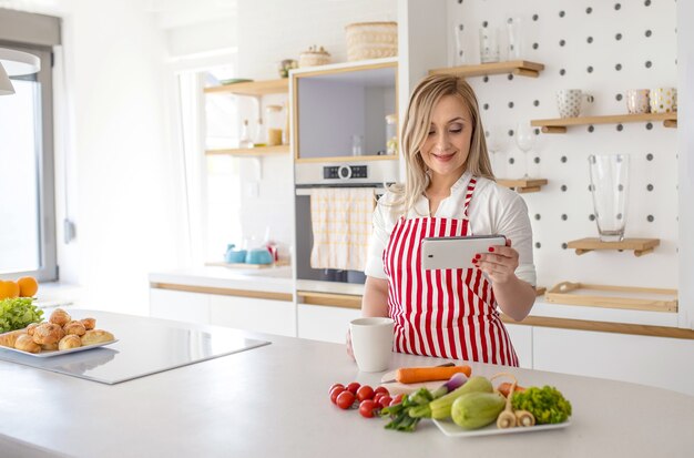 Free photo young caucasian female with red striped apron holding mug looking at recipe on phone in kitchen