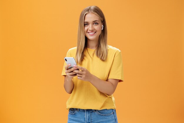 Young caucasian girl with tanned skin and fair hair using wireless earphones to call friend via smartphone holding cellphone against chest, smiling cheerfully at camera getting used to new technology