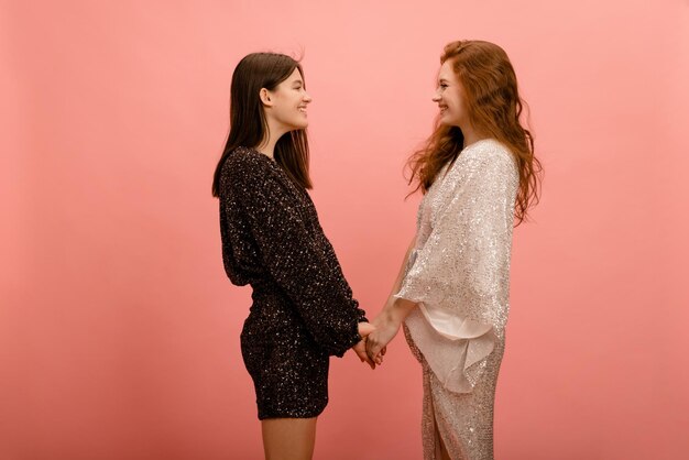 Young caucasian girls in shiny dresses smiling look at each other and hold hands on pink background Women's friendship concept