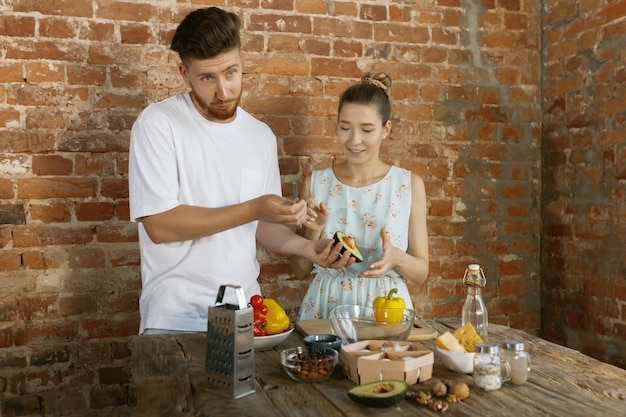 Young caucasian happy couple cooking together using vegetables, cheese, eggs and nuts in recipe against brick wall in their kitchen. Nutrition, healthy food, family, relations, domestic life concept.