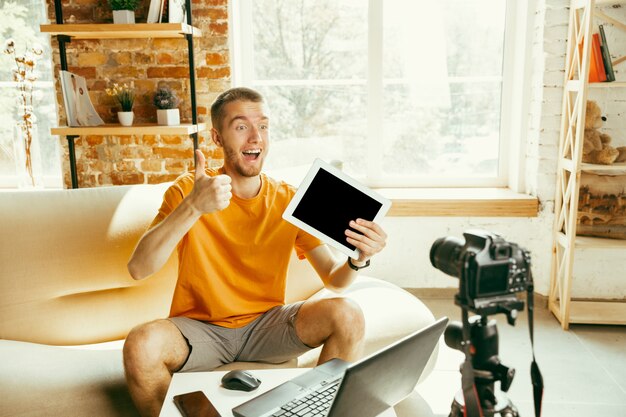 Young caucasian male blogger with professional camera recording video review of tablet at home. Blogging, videoblog, vlogging. Man making vlog or live stream about photo or technical novelty.