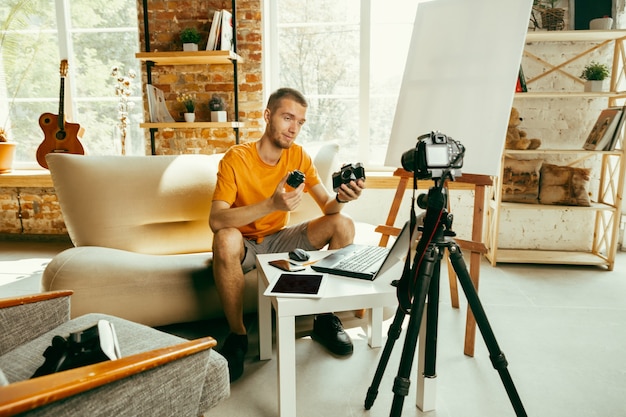 Young caucasian male blogger with professional equipment recording video review of camera at home. Blogging, videoblog, vlogging. Man making vlog or live stream about photo or technical novelty.