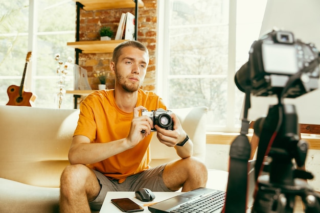 Free photo young caucasian male blogger with professional equipment recording video review of camera at home. blogging, videoblog, vlogging. man making vlog or live stream about photo or technical novelty.