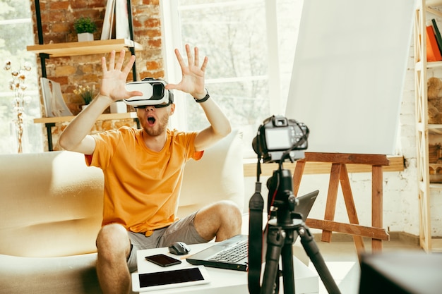 Young caucasian male blogger with professional equipment recording video review of VR glasses at home