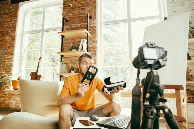 Young caucasian male blogger with professional equipment recording video review of VR glasses at home