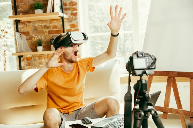 Young caucasian male blogger with professional equipment recording video review of VR glasses at home.