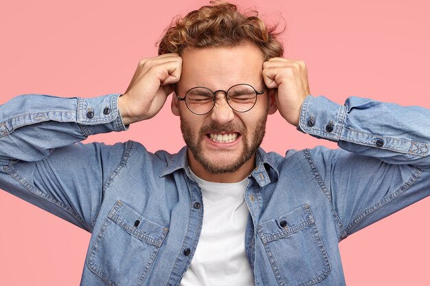 Young Caucasian male suffers from headache and migraine, keeps fists on temples, clenches teeth, has frustrated expression, dressed in denim stylish shirt, isolated over pink wall. Sick guy