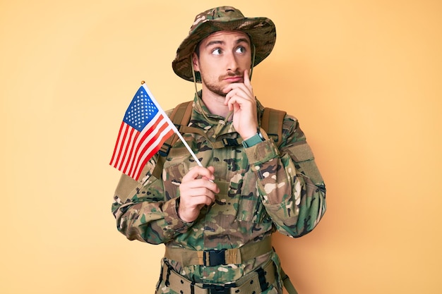 Free photo young caucasian man wearing camouflage army uniform holding usa flag serious face thinking about question with hand on chin thoughtful about confusing idea