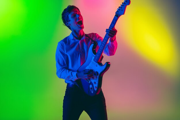 Young caucasian musician, guitarist playing on gradient space in neon light. Concept of music, hobby, festival