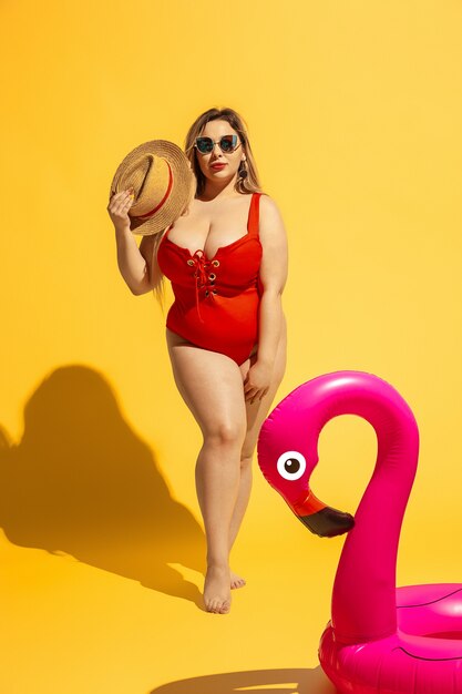 Young caucasian plus size female model's preparing vacations on yellow wall. Woman in red swimsuit and hat posing with swimring.