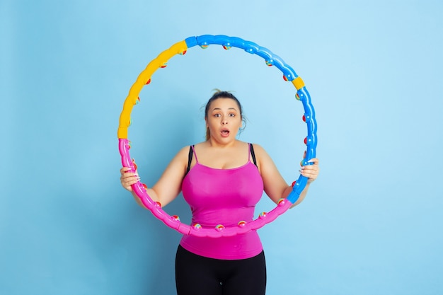 Young caucasian plus size female model's training on blue background. Concept of sport, human emotions, expression, healthy lifestyle, body positive, equality. Astonished holding the hoop, shocked.