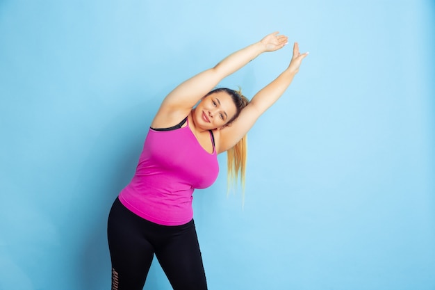 Free photo young caucasian plus size female model's training on blue background. concept of sport, human emotions, expression, healthy lifestyle, body positive, equality. making stretching exercises.