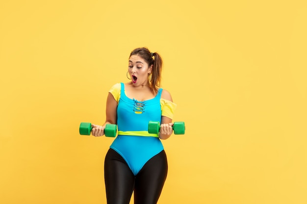 Free photo young caucasian plus size female model's training on yellow wall. copyspace. concept of sport, healthy lifestyle, body positive, fashion, style. stylish woman emotional practicing with weights.