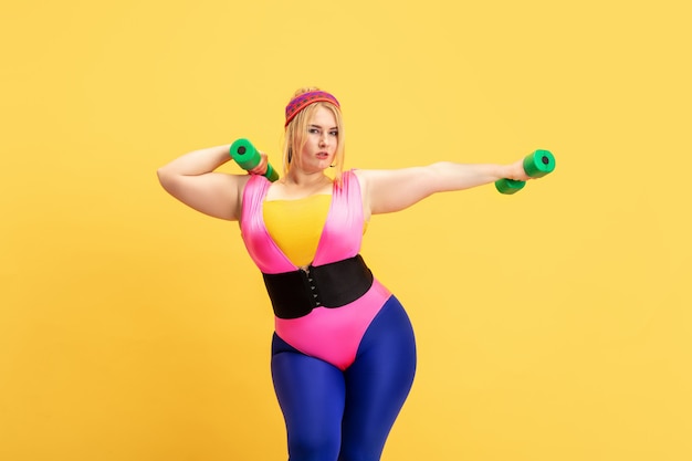 Free photo young caucasian plus size female model's training on yellow wall. copyspace. concept of sport, healthy lifestyle, body positive, fashion, style. stylish woman practicing with green weights.