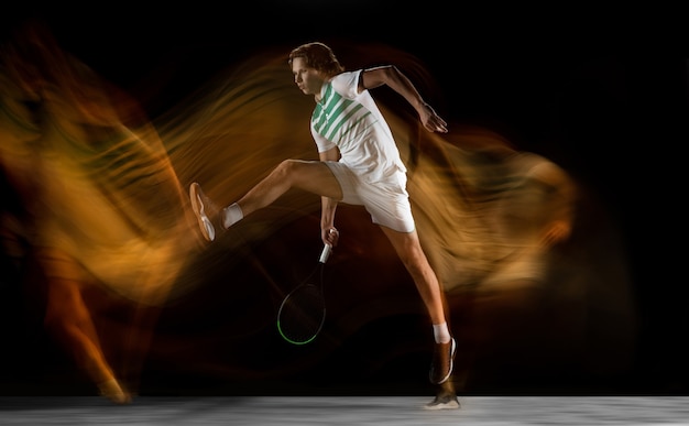 Young caucasian professional sportsman playing tennis on black wall in mixed light