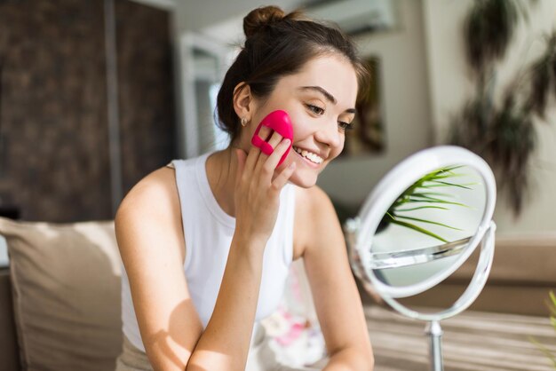 A young caucasian smiling woman is doing a facial massage looking in the mirror