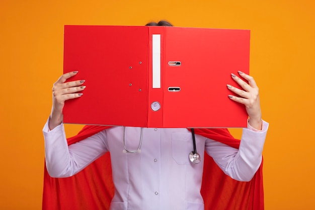 Free photo young caucasian superhero girl in red cape wearing doctor uniform and stethoscope with glasses holding open folder in front of face