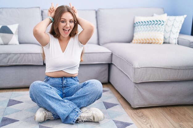 Young caucasian woman sitting on the floor at the living room posing funny and crazy with fingers on head as bunny ears smiling cheerful