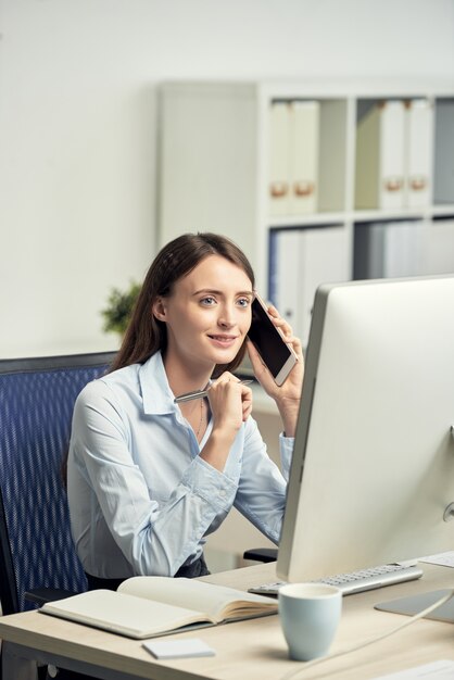 Young Caucasian woman sitting in office in front of computer screen and talking on phone