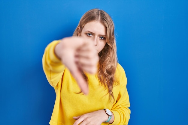 Young caucasian woman standing over blue background looking unhappy and angry showing rejection and negative with thumbs down gesture bad expression