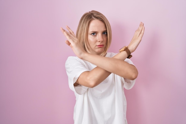 Free photo young caucasian woman standing over pink background rejection expression crossing arms doing negative sign, angry face