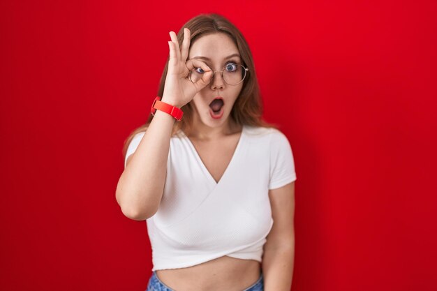 Young caucasian woman standing over red background doing ok gesture shocked with surprised face eye looking through fingers unbelieving expression