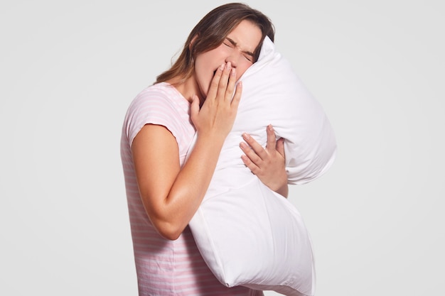 young Caucasian woman wants to sleep, keeps hand on mouth, dressed in casual clothes, holds pillow, has tired expression, isolated on white. Morning and awakening concept