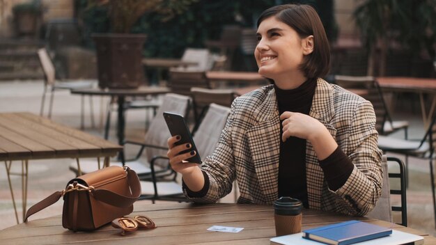 Young charming businesswoman looking happy using smartphone on coffee break in cozy courtyard of cafe