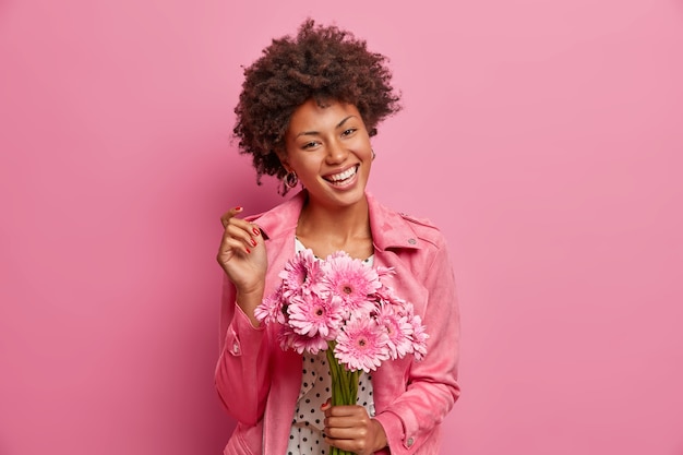 Young cheerful African American woman with natural makeup, broad smile, holds bouquet of flowers, going to congratulate friend, enjoys pleasant smell of gerberas