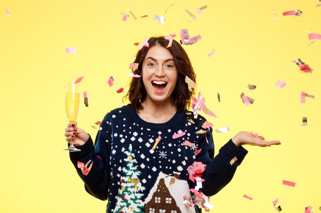 Young cheerful beautiful brunette girl in cosy knited sweater smiling holding glass of champagne over yellow background with falling confetti.