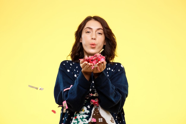 Free photo young cheerful beautiful brunette girl in cosy knited sweater smiling with handful of confetti over yellow background.