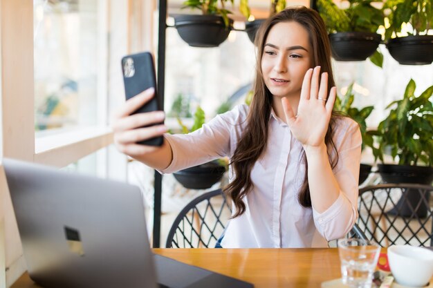 Young cheerful woman posing while photographing herself on smart phone for a chat with her friends, attractive smiling hipster girl making self portrait on cell telephone while sitting in cafe