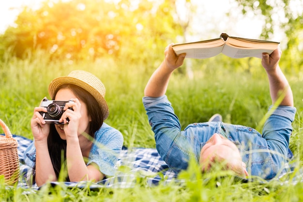 Young cheerful woman taking photo and man reading book in grass