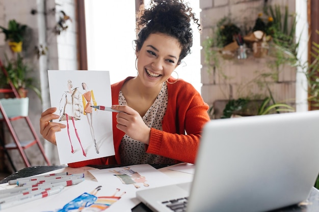 Young cheerful woman with dark curly hair sitting at the table happily showing fashion illustration in laptop while spending time in modern cozy workshop with big windows