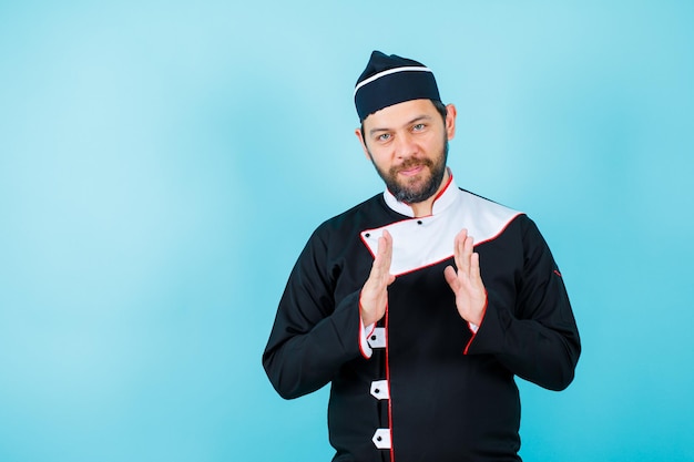 Young chef is looking at camera by holding up his hands on blue background