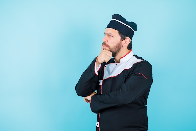 Young chef is thinking by looking up and putting hand on chin on blue background