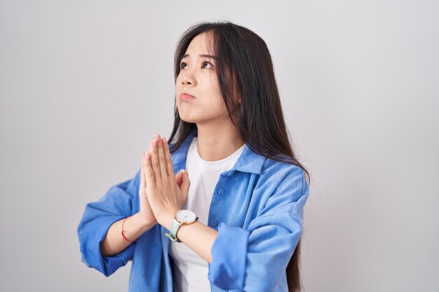 Young chinese woman standing over white background begging and praying with hands together with hope expression on face very emotional and worried. begging.