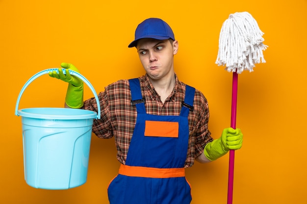 Free photo young cleaning guy wearing uniform and cap with gloves holding mop and bucket isolated on orange wall